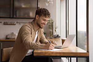 A man sitting at a desk in a kitchen writing on paper with a laptop on the desk