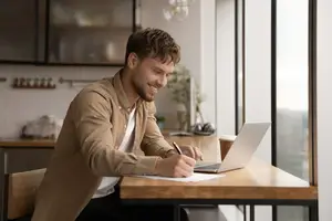 A man sitting at a desk in a kitchen writing on paper with a laptop on the desk