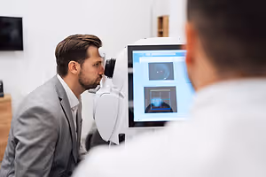 An adult man wearing a gray suit and a white shirt is looking at a computer monitor while wearing a device on his head, possibly undergoing an eye examination in a clinic or hospital.