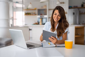 Woman sitting at a table in a kitchen with a laptop and tablet smiling at the camera