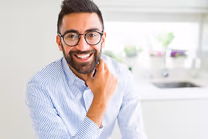 A man with a beard wearing glasses and a blue striped shirt is smiling in a kitchen with a sink and faucet behind him.