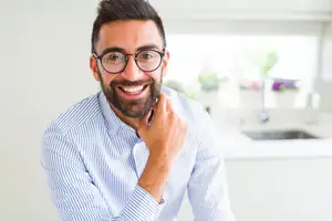 A man with a beard wearing glasses and a blue striped shirt is smiling in a kitchen with a sink and faucet behind him.