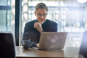 A smiling man sitting at a desk working on a laptop with a coffee mug nearby.