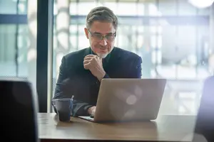 A smiling man sitting at a desk working on a laptop with a coffee mug nearby.