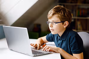 A young boy wearing glasses is using a laptop while sitting on a chair in a room with a shelf and a desk.