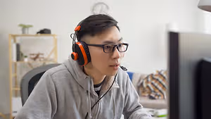 A man sitting at a desk with a computer monitor wearing headphones and glasses, possibly playing a video game