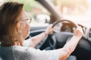 An older woman is driving a car on a sunny day, wearing glasses, and holding the steering wheel with both hands.