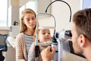 A young girl with her mother is having her eyes examined by a male optometrist using an eye exam device.