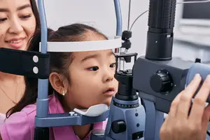 A girl with a headband looks through an ophthalmoscope while a woman smiles behind her in a medical setting.