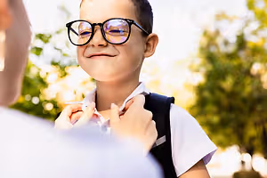 A boy wearing glasses and a backpack smiles while a person adjusts his collar in an outdoor setting.