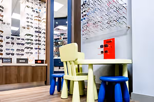 A kid's table and chairs are placed in a shop, with many glasses displayed on the wall and a sign that reads 'HUGO' on the wall. The shop has a blue door and a wooden counter. The shop has a wooden floor and a white wall. The shop has a bright light on the ceiling.