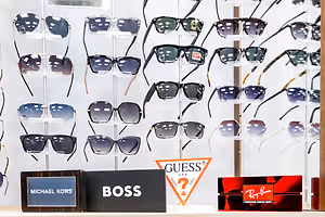 A wall display of sunglasses from various brands, including Guess, Michael Kors, and Ray-Ban, with each pair neatly arranged in a row on transparent stands, showcasing their unique designs and colors. The backdrop is white, highlighting the contrast of the sunglasses.