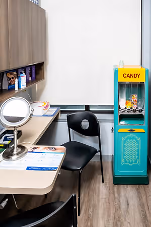 A room with a desk and chair, a candy machine, and a mirror. There are also cabinets with various items on them, and a window on the wall.