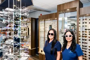 Two women in blue uniforms stand in an optical shop, smiling and wearing white sunglasses, with a display of glasses behind them.