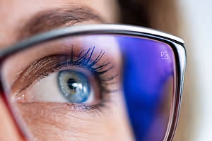 A close-up of a woman's eye with blue iris and eyelashes reflected in her glasses.