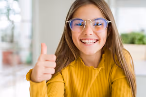 A girl with glasses and a yellow sweater is smiling and giving a thumbs up in a room with a blurry background