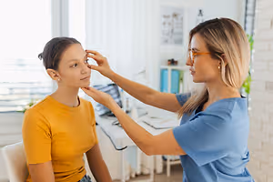 A woman sitting in a chair while a woman stands next to her and adjusts her glasses