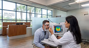A woman in a white lab coat operates a retinal imaging device on a man wearing a blue shirt in an Orlando Optometry Group office.