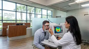 A woman in a white lab coat operates a retinal imaging device on a man wearing a blue shirt in an Orlando Optometry Group office.