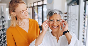 Two women smiling and standing next to each other in a store, one of them trying on glasses