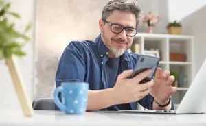 A man sits at a desk with a laptop, holding a phone, and wearing glasses.