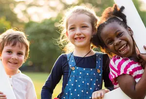 Three children, two girls and a boy, smiling and holding onto a white structure while posing for a photograph in a park.