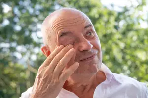 An older man is smiling and touching his eye with his hand