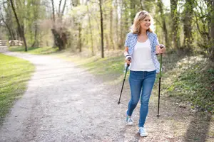 A woman walking on a dirt path in a forest with a smile on her face and holding a pair of trekking poles
