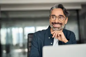 A man with a beard and glasses smiles while looking at a laptop screen inside an office building