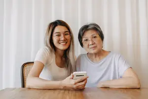 A woman wearing glasses and holding a white phone and an older woman sitting next to her wearing eyeglasses on a wooden table in front of a white curtain