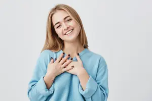 A woman with blonde hair is smiling and posing for a photo while wearing a light blue long-sleeved shirt and holding her chest with both hands.