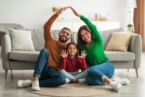 A family with a man, a woman, and a girl sitting on the floor in front of a couch, all of them smiling and posing for a photo
