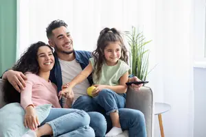 A family of four sitting on a couch, two adults and two children, with the girl holding a remote and an apple, and the boy smiling.