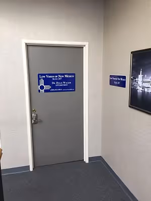 A sign on the door of Low Vision of New Mexico Suite 207 with a photo of a city on the wall and a gray carpeted floor.
