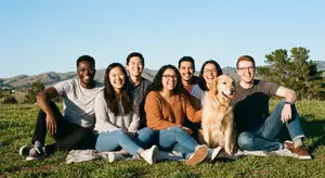 A group of people are sitting on a blanket in a grassy field with a dog. There are hills in the background and a blue sky.