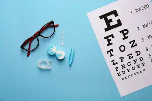 A blue surface displays a pair of glasses, an eye chart, a pair of scissors, a contact lens, a pen, and a white tape