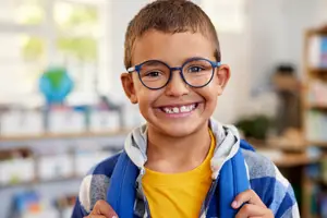 boy wearing glasses and a backpack smiling in a classroom