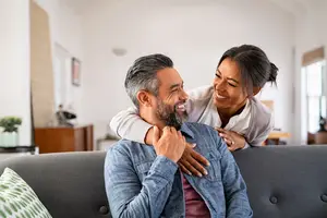 A man with gray hair and a beard is sitting on a couch with a smiling woman standing behind him. The woman is placing her hands on the man's shoulders. They are both looking at something with a smile on their faces. Behind them is a wooden cabinet with a potted plant on top. There is a picture frame on the wall near the window.