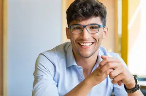 A smiling young man with glasses wearing a light blue button up shirt sits at a table.