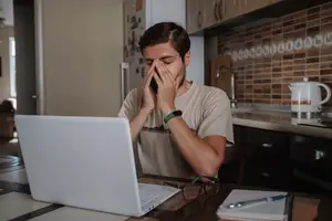 A man sitting at a table in the kitchen with his hands on his face, looking tired and stressed