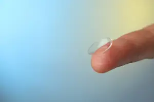 A finger holding a clear contact lens against a blue and yellow background