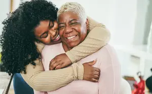 A woman with curly hair is smiling and hugging an older woman with white hair in a living room