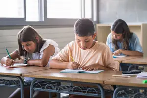 Three children sitting at desks in a classroom, writing with pencils