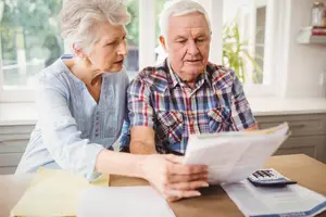 An elderly couple reviewing documents at a table, including a calculator and papers.
