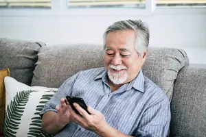 A man is sitting on a couch and looking at his cell phone. He is smiling and seems to be enjoying what he is looking at. He has gray hair and a white beard. He is wearing a blue polo shirt. There are pillows on the couch. Behind him is a glass window with blinds. The room is well lit.