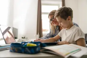 A boy and a woman are working on a laptop with books and a pen on a desk.