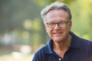 A man with gray hair wearing glasses and a blue polo shirt is smiling and looking forward in a green park. 