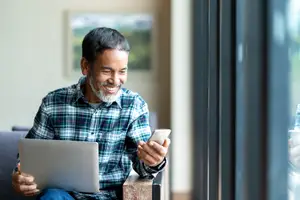 An older man sitting on a couch, smiling and holding a cell phone and laptop