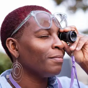 A woman with red hair is looking through a telescope while wearing glasses and earrings.