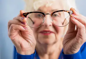 A senior woman with gray hair wearing a blue shirt adjusting her eyeglasses.